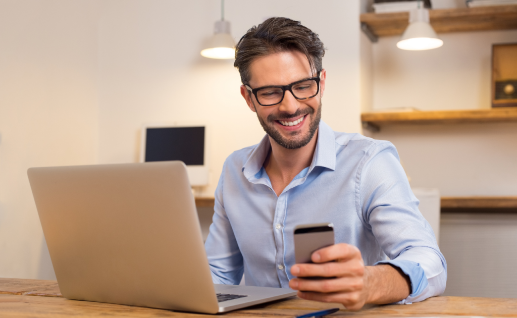 Mann mit einem Handy vorm Laptop